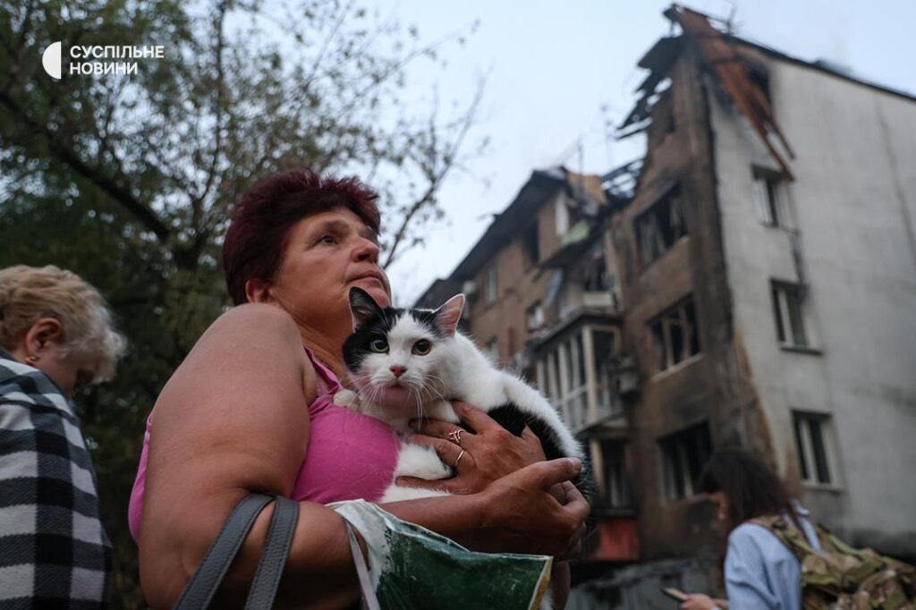 russia strikes kyiv 10 hours—two women killed including 22-year-old metro police officer woman holds cat front residential building damaged russian shahed drone 2025 people watch burn after attack suspilne news